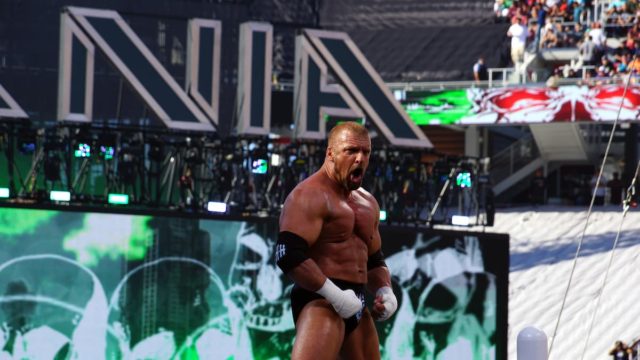 Triple H standing in a WWE ring, wearing a suit and holding a microphone – symbolising his wrestling and executive careers