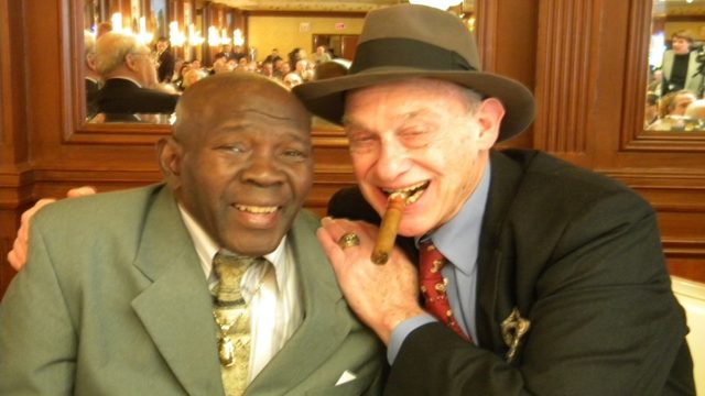 Bert Sugar wearing his trademark fedora and cigar while ringside at a boxing event.
